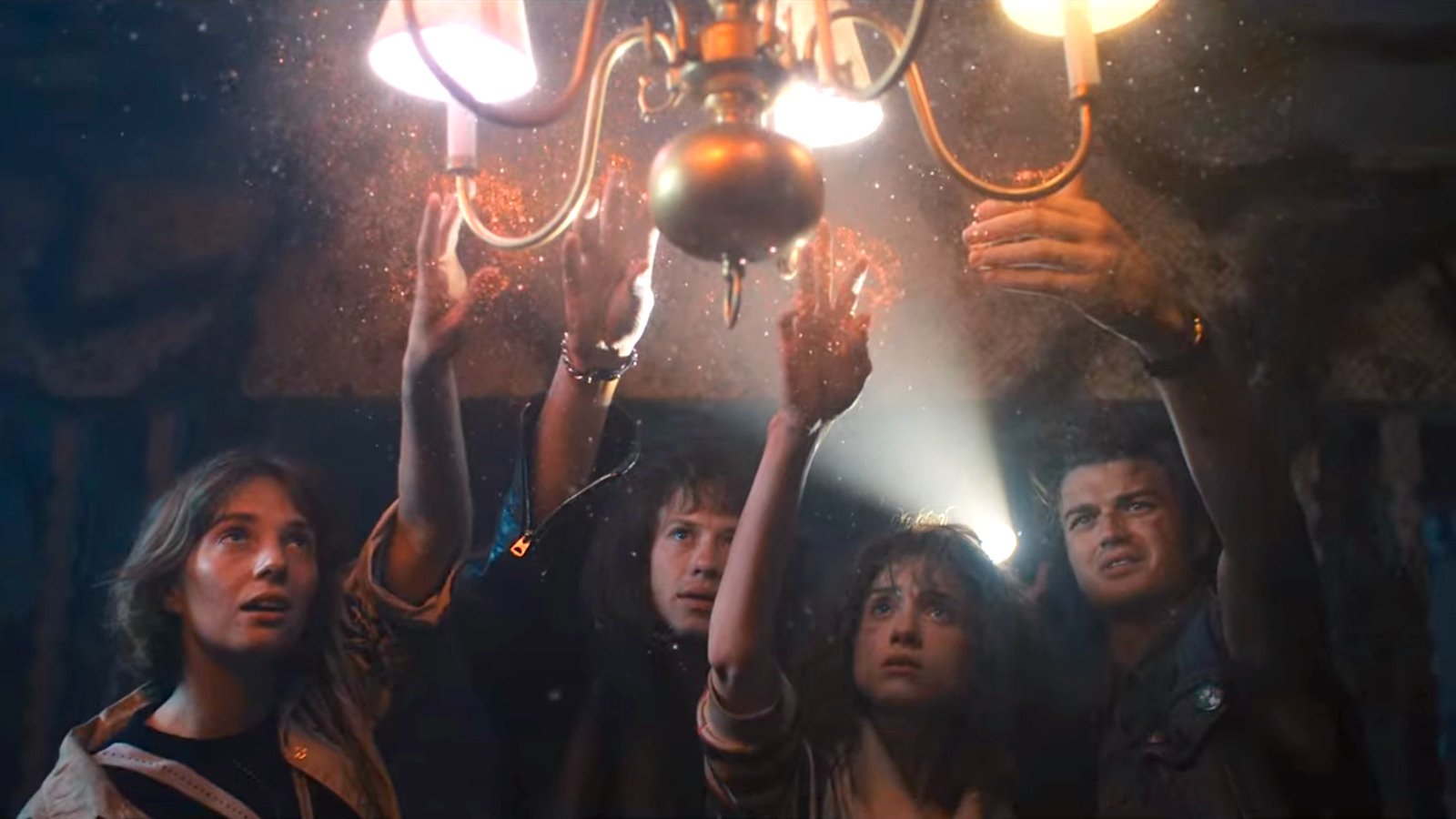 Robin, Eddie, Nancy, and Mike from 'Stranger Things' reaching to touch a lit chandelier with sparkly dust emanating from it 