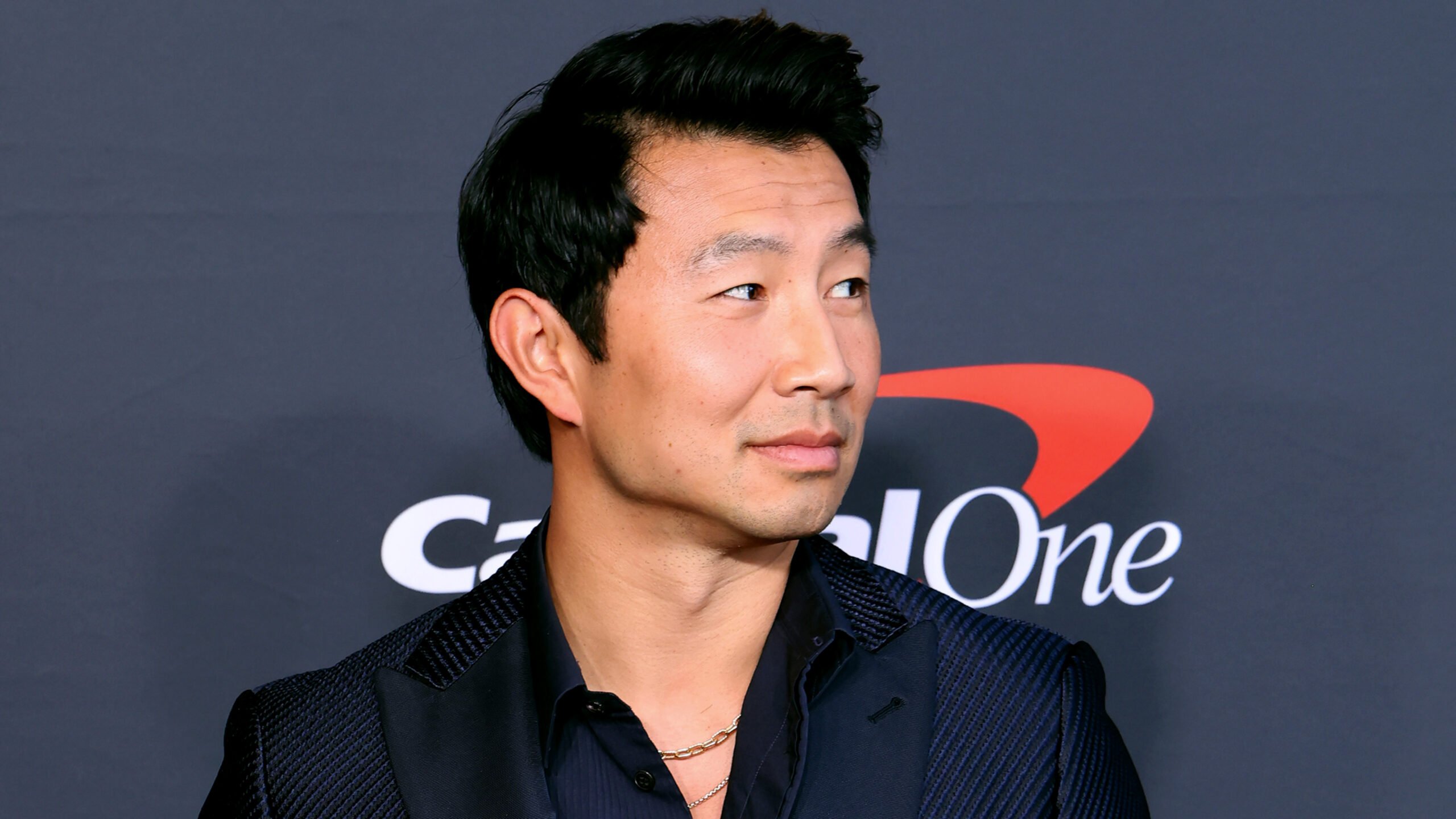 Simu Liu in a shoulders-up portrait in dark shirt and jacket, smiling slightly and looking off-camera