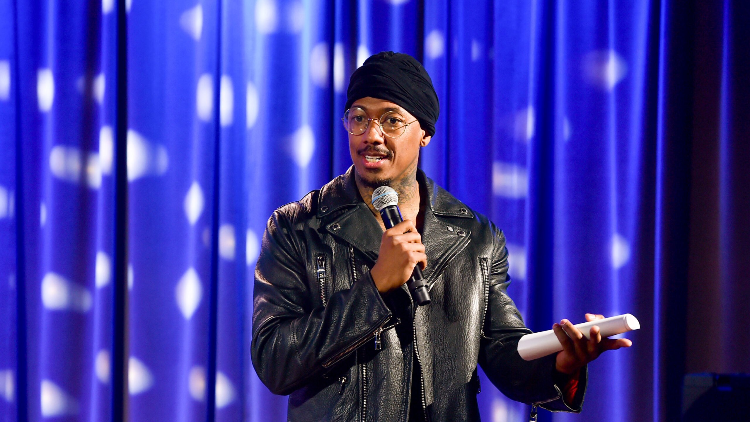 Nick Cannon is shown from the waist up, wearing a black motorcycle jacket, wire rimmed glasses and a black beanie, speaking into a microphone