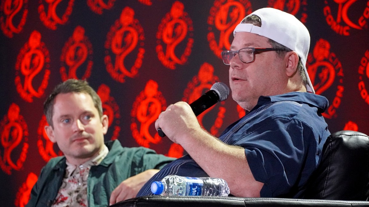 Sean Astin and Elijah Wood give a talk on a stage with a red background.