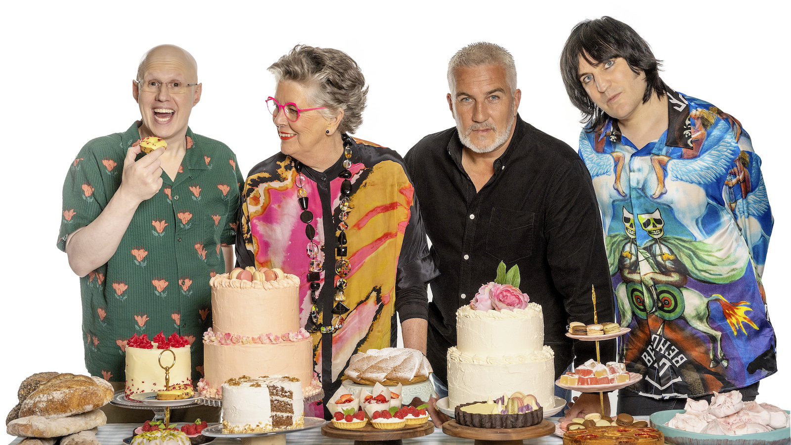 The hosts of The Great British Baking Show stand behind a table full of baked goods