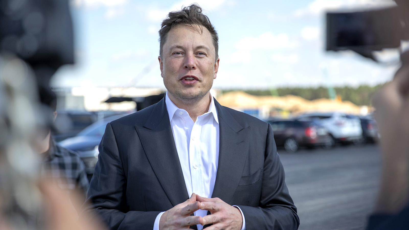 Elon Musk in a black and white suit speaking to a barrage of camera pointed at him