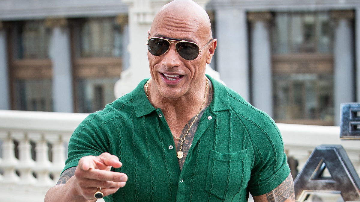 Dwayne Johnson is in a green shirt with black sunglasses and is gesturing.