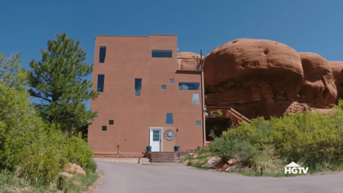 The exterior of a plain building perched beside a huge red rock. 