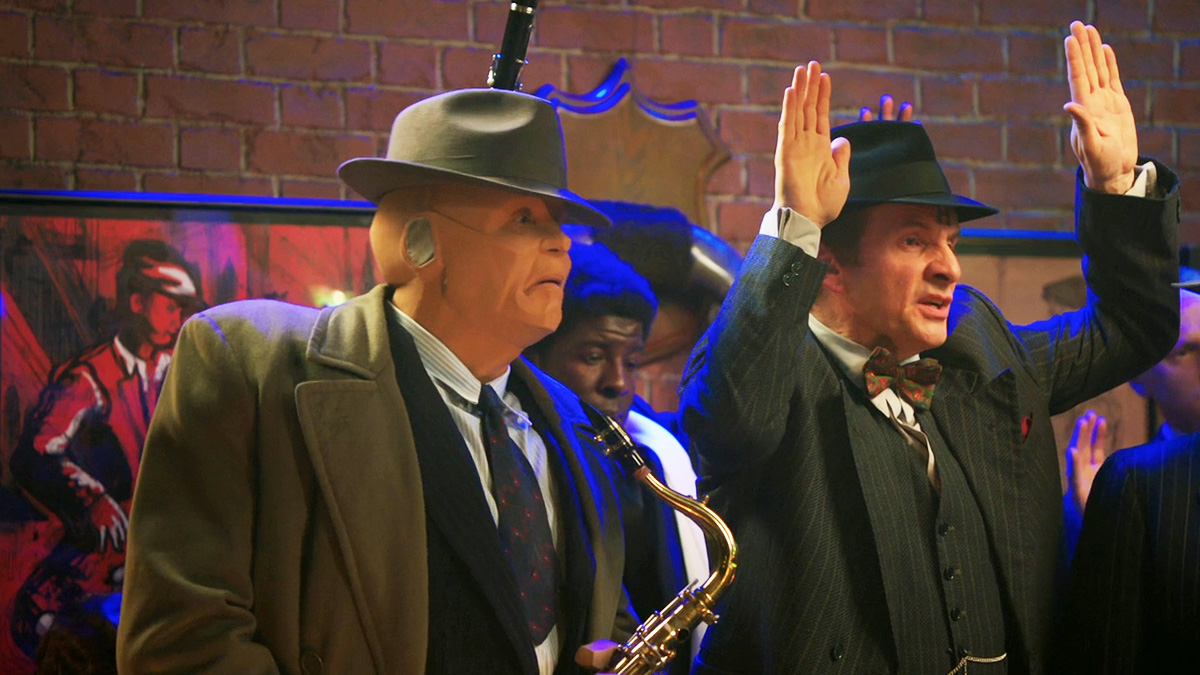 Kryten holds a saxophone as Rimmer stands next to him, raising his hands. They're both dressed in fedoras and trench coats from olden times.