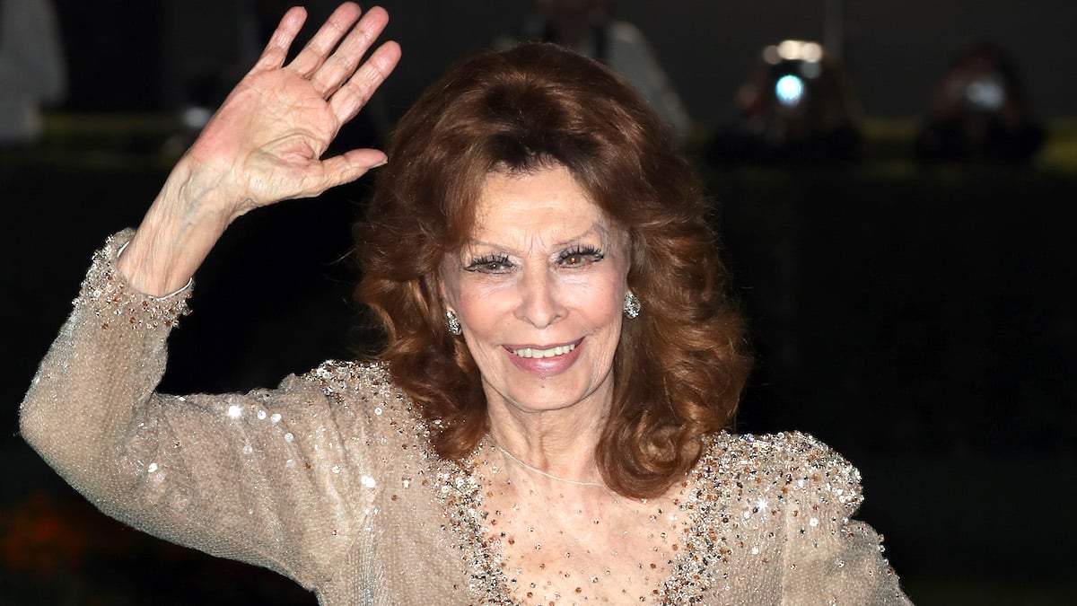 Sophia Loren in a frilled gold dress with a net plunge smiling and waving