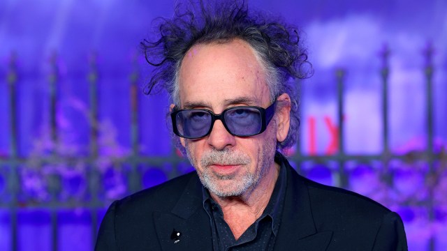 Tim Burton strikes a pose wearing blue tinted glasses. He appears unshaven with disheveled hair at the premiere of 'Wednesday,' a purple backdrop with an ominous metal fence behind him.