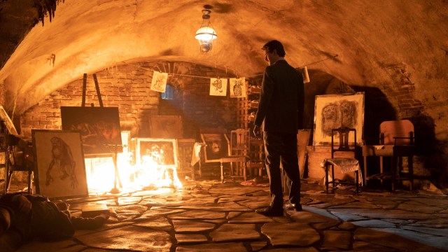 Man stands in cellar, in front of paintings on fire.